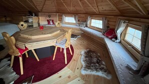 A cozy cabin interior with a wooden dining table, chairs, a fur rug, and a window with curtains. - Finnish Kota with Jacuzzi, sauna, heated swimming pool and wellness treatments (Voissant)