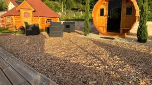A wooden cabin with a red roof, a round wooden structure with a door, and a small garden area with mulch and a bench. - Finnish Kota with Jacuzzi, sauna, heated swimming pool and wellness treatments (Voissant)