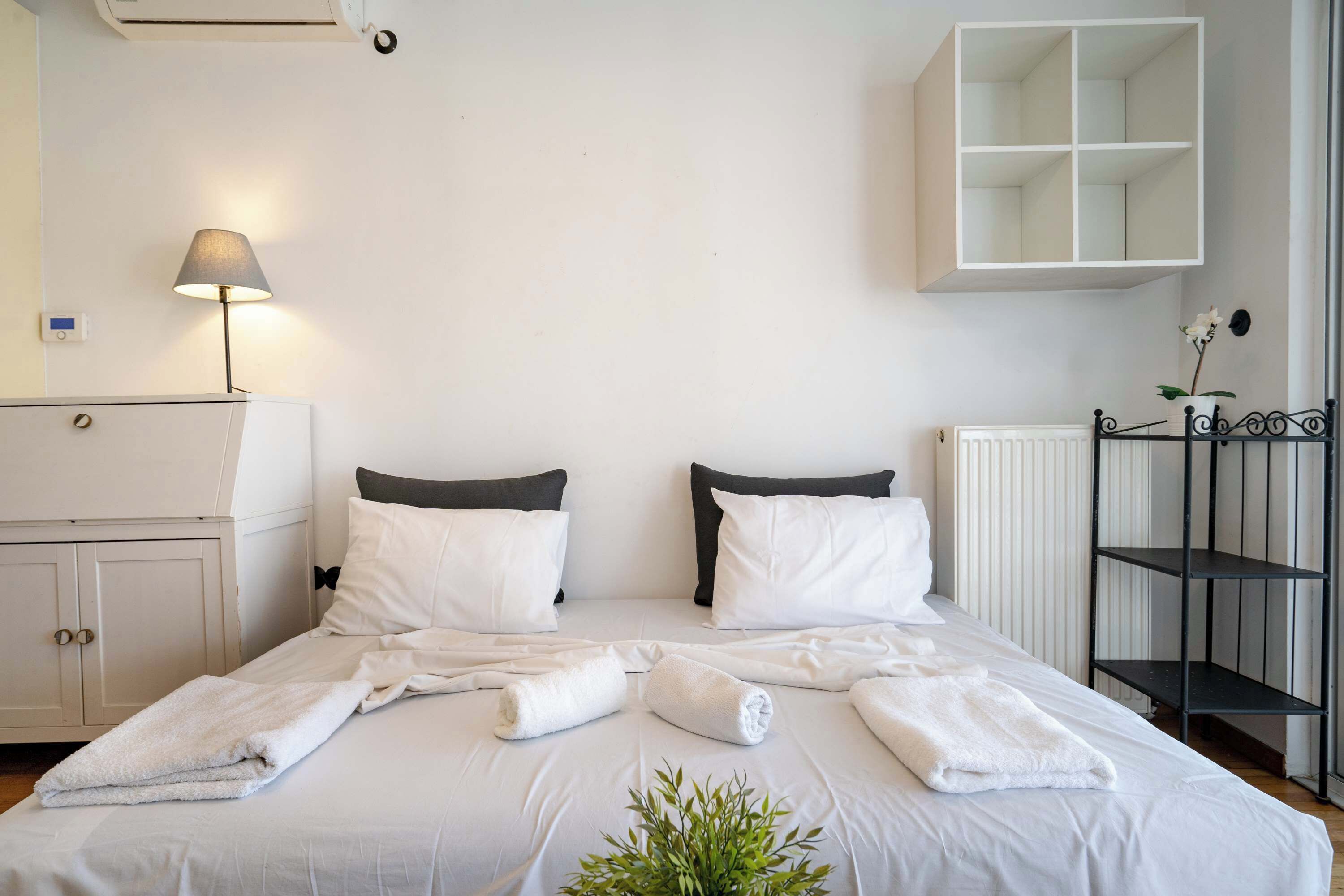 Un lit bien fait, avec du linge de lit et des serviettes blanches, une lampe de chevet, une armoire blanche et un radiateur.