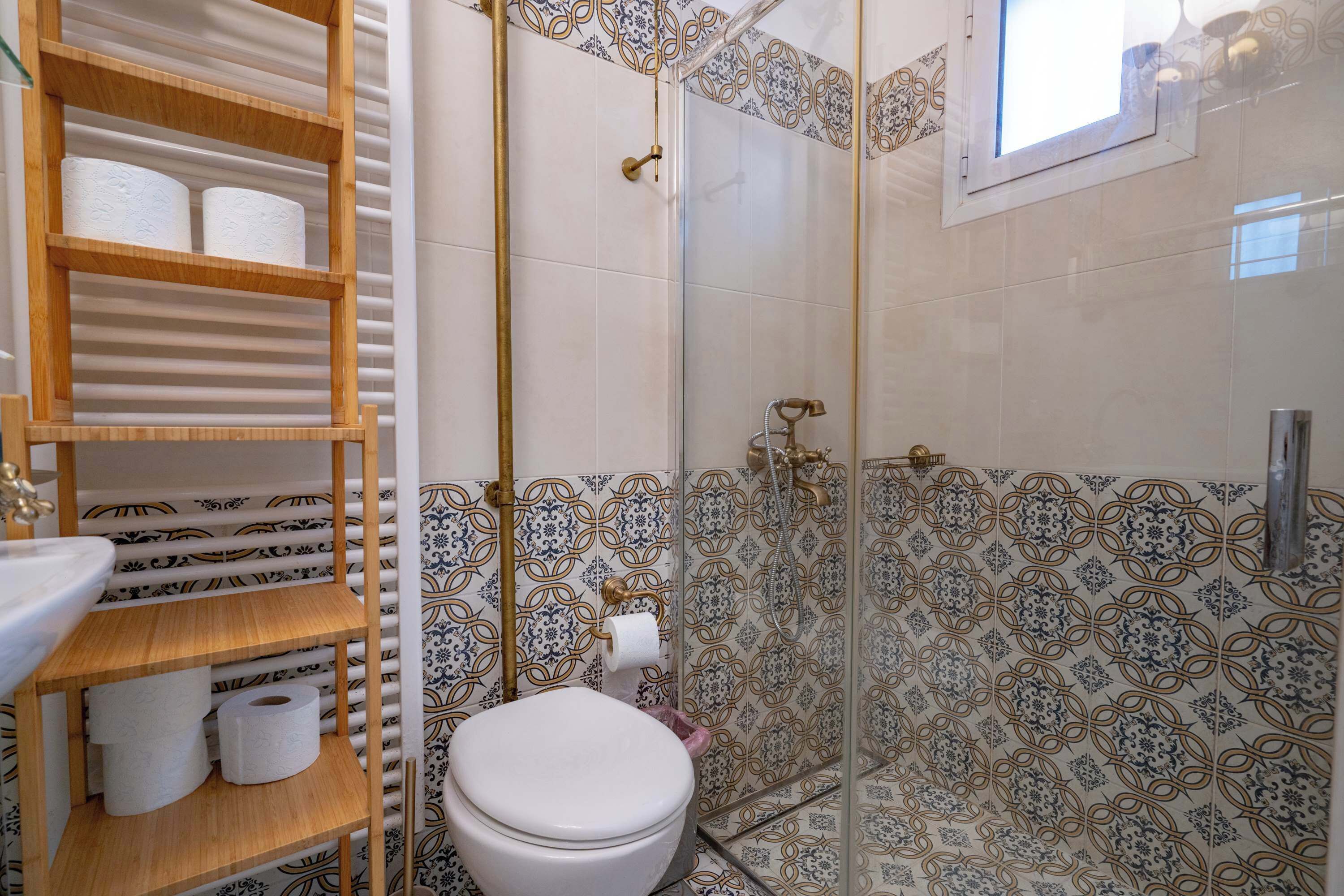 Un baño con inodoro, repisas con papel higiénico, ducha con puerta de vidrio y azulejos estampados.