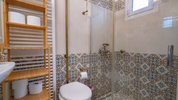 Un baño con inodoro, repisas con papel higiénico, ducha con puerta de vidrio y azulejos estampados.