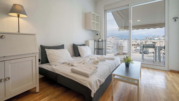 A modern bedroom with a large bed, a bedside table, a lamp, and a view of the city through large windows. - City Panorama Studio by ΜΡS (Athens)