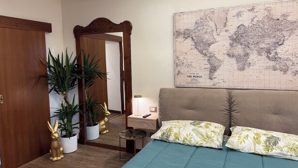 A bedroom with a large world map on the wall, a bed with green and white patterned pillows, a wooden door, and potted plants. - IRIDE HOME _ Wellness & Relaxation (Bergamo)