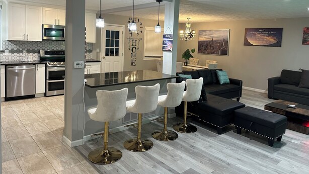 A modern kitchen with stainless steel appliances, a central island with bar stools, and a dining area with a black sofa and ottoman. - Luxurious 5-bedroom house with Game Room in Stone Mountain (Stone Mountain)
