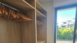 Un dressing accessible sans porte, doté d’étagères et de tiroirs en bois, un banc avec des serviettes, et une fenêtre offrant une vue sur les bâtiments et la verdure.