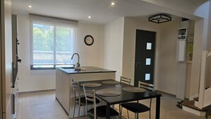 Une cuisine moderne dotée d’un îlot central, d’une table à manger avec des chaises, d’une fenêtre avec des stores et d’une porte avec un panneau en verre dépoli.