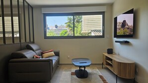 Un salon moderne avec un canapé, une petite table, un téléviseur et une fenêtre donnant sur des immeubles et des arbres.