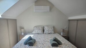 Une chambre avec un plafond en pente, un lit avec une literie à motifs floraux, deux oreillers, deux lampes de chevet et un climatiseur fixé au mur.
