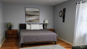 Ein Schlafzimmer mit Bett, Nachttisch, Lampe, einer Uhr, einer Pflanze und einem Sessel.