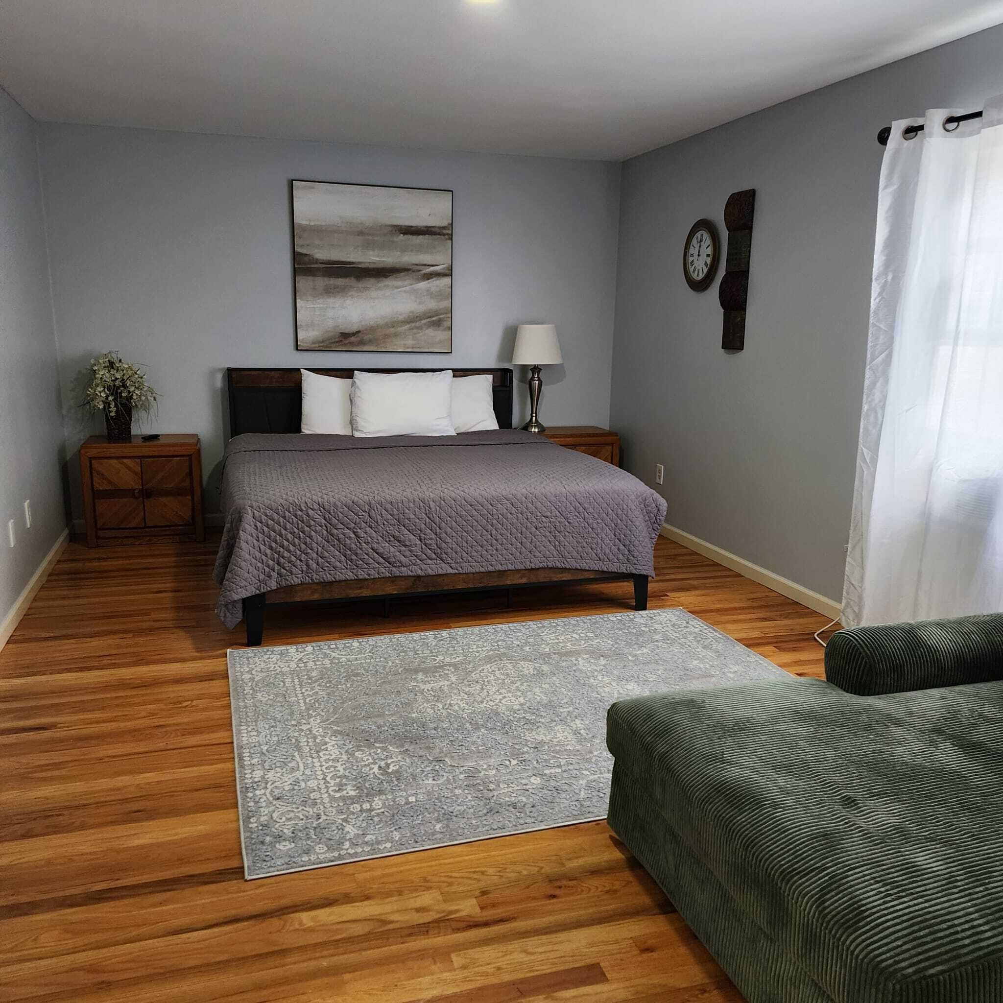 Quarto com cama, mesa de cabeceira, abajur, relógio, planta e cadeira.