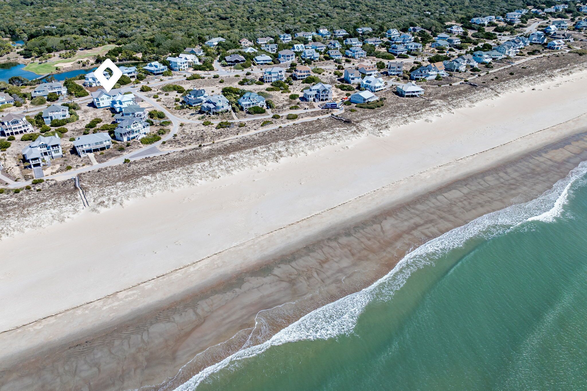 An aerial view of a coastal residential area with houses along the beach, a clear view of the ocean, and a small body of water in the background.