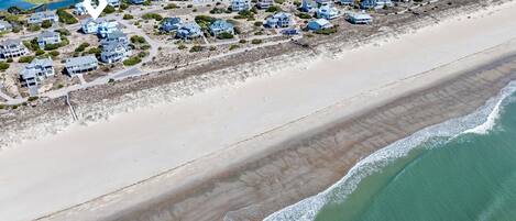 An aerial view of a coastal residential area with houses along the beach, a clear view of the ocean, and a small body of water in the background.