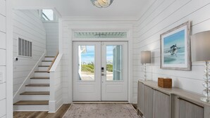 A hallway with a wooden staircase, a double door with glass panels, a patterned rug, a console table with a lamp, and a framed picture on the wall. - Steps to the Beach: 6BR Gem / Ocean Views / BHI + Shoals Club / 2 Carts (Bald Head Island)