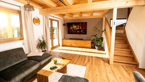 A modern living room with a wooden ceiling, a large TV, a sofa, a coffee table, and a staircase. - Heimatzeit - Chalet with whirlpool and sauna (Reichenbach)