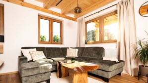 A modern living room with a grey sectional sofa, wooden coffee table, and large windows with curtains. - Heimatzeit - Chalet with whirlpool and sauna (Reichenbach)
