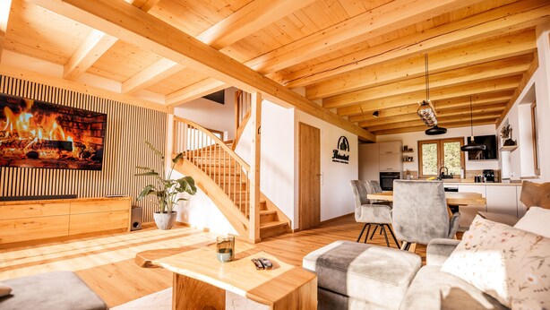 A spacious living area with a wooden ceiling, a fireplace, a staircase, and a kitchen in the background. - Heimatzeit - Chalet with whirlpool and sauna (Reichenbach)