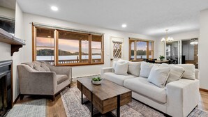 A living room with a fireplace, a sofa, a coffee table, and large windows offering a view of a lake. - Lakefront Cottage with Stunning Sunset Views at Indian Lake! (Belle Center)