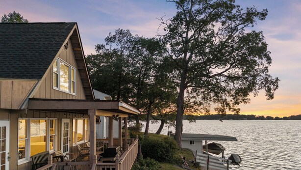 A wooden house with a deck overlooking a lake, a covered dock, and a large tree in the background. - Lakefront Cottage with Stunning Sunset Views at Indian Lake! (Belle Center)