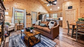 Un interior acogedor de cabaña con techo de madera, una sala de estar con sofá y mesa de centro, una zona de cocina y una mesa de comedor.