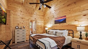 Un dormitorio tipo cabaña de madera con cama, mesita de noche, lámpara y un cuadro en la pared.