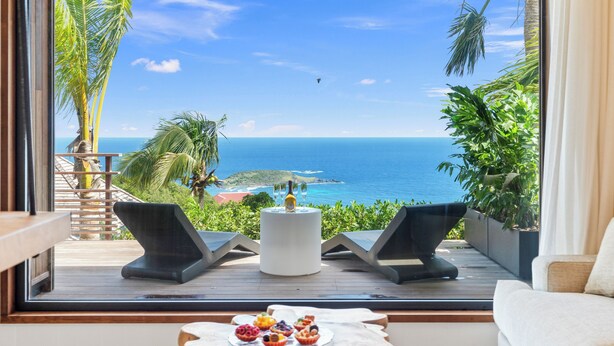 A balcony with a view of the ocean, two lounge chairs, a table with a bottle and glasses, and a small table with pastries. - Lil' Home (St. Barthelemy)