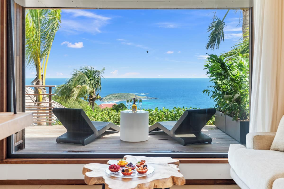 A balcony with a view of the ocean, two lounge chairs, a table with a bottle and glasses, and a small table with pastries.