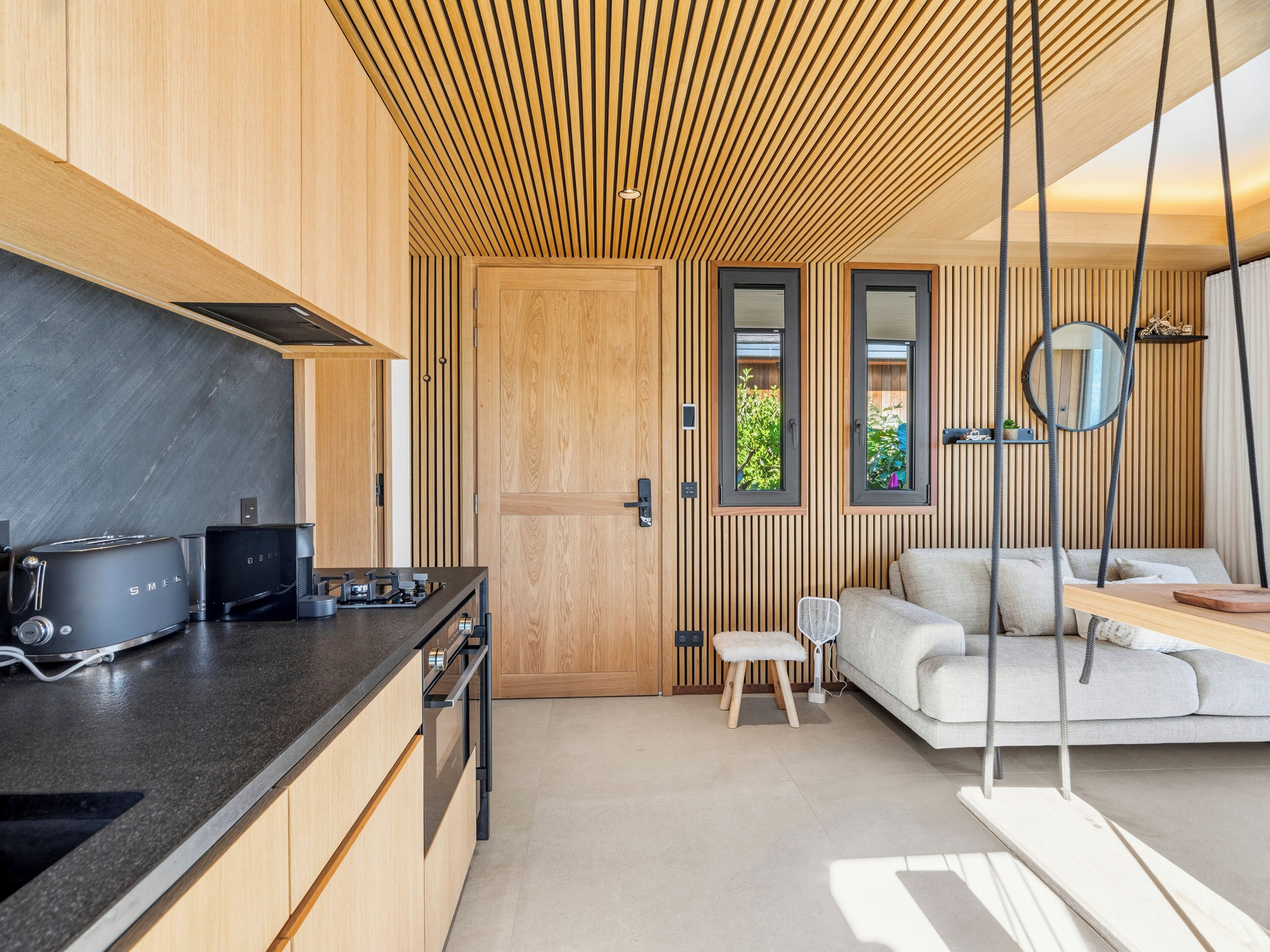 A modern kitchen with wooden cabinetry and a black countertop. There is a small seating area with a white sofa and a wooden table.