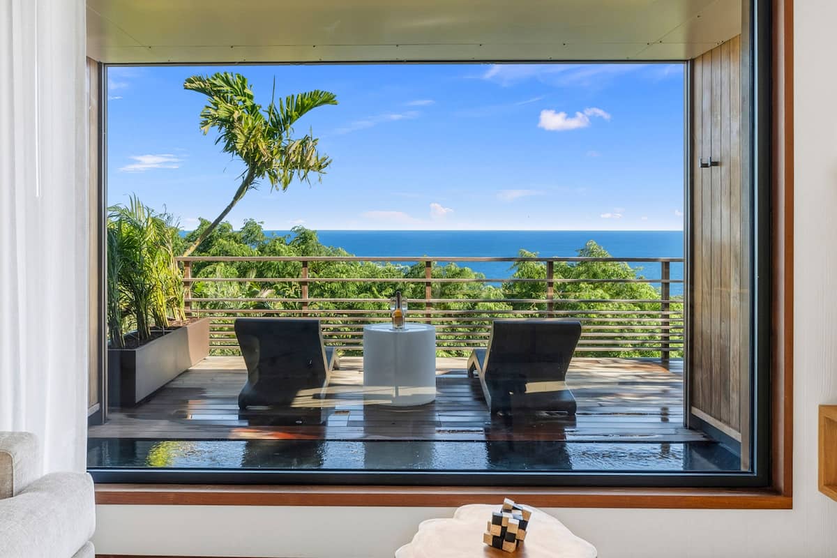 A room with a large window overlooking a balcony with two chairs and a table, a small wooden table with a Rubik's cube, and a view of the ocean and greenery.