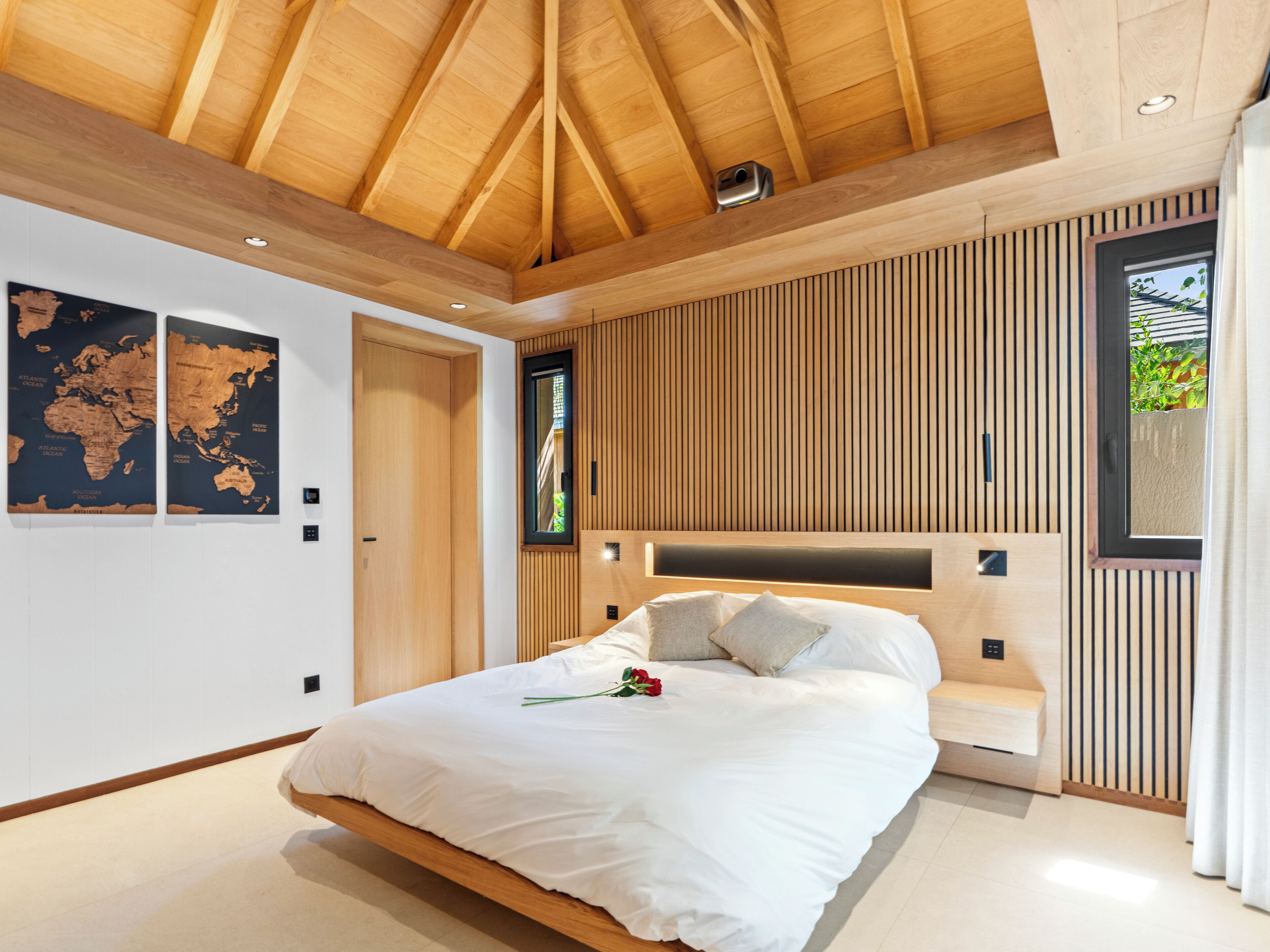A modern bedroom with a wooden ceiling, a large bed, a world map on the wall, and a window with a view of greenery.