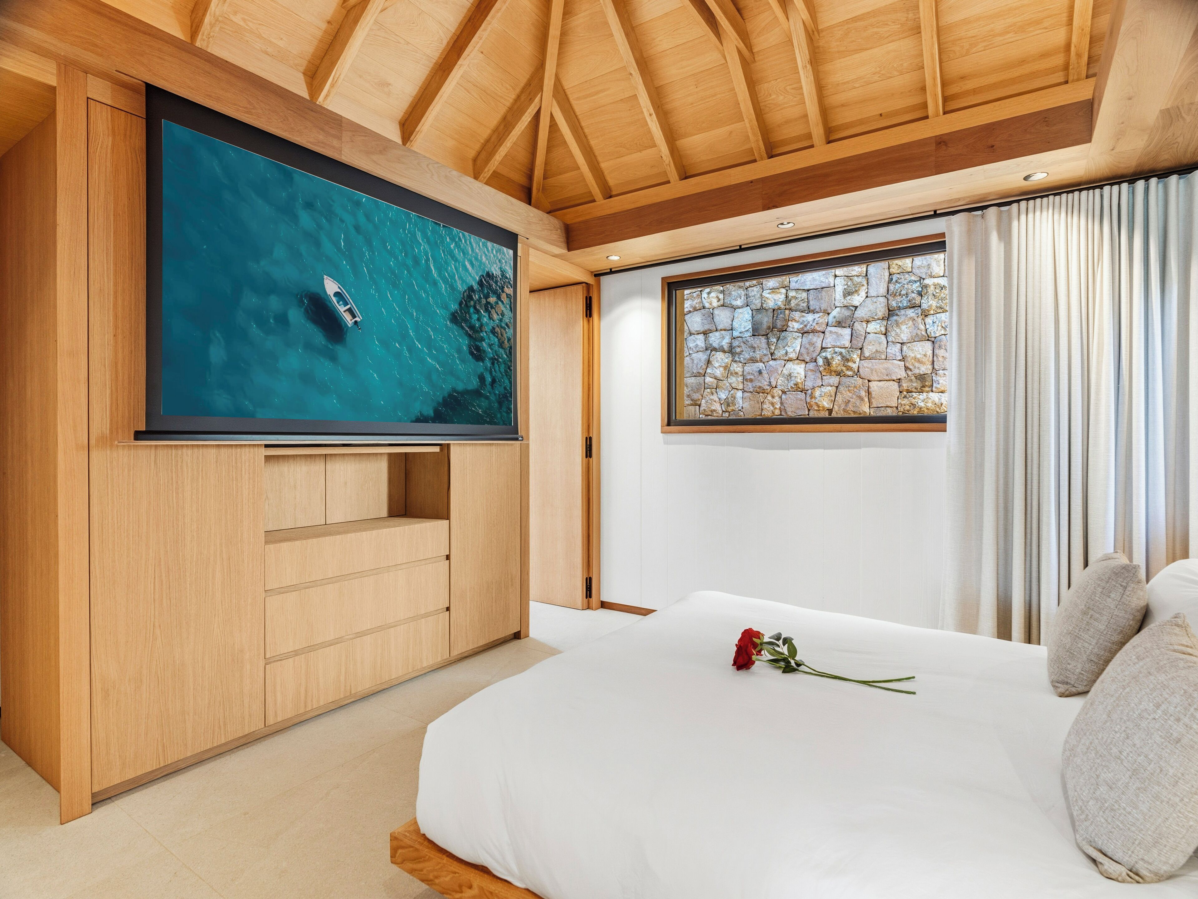 Quarto com televisão grande, teto de madeira, parede de pedra e cama com almofadas e uma rosa.