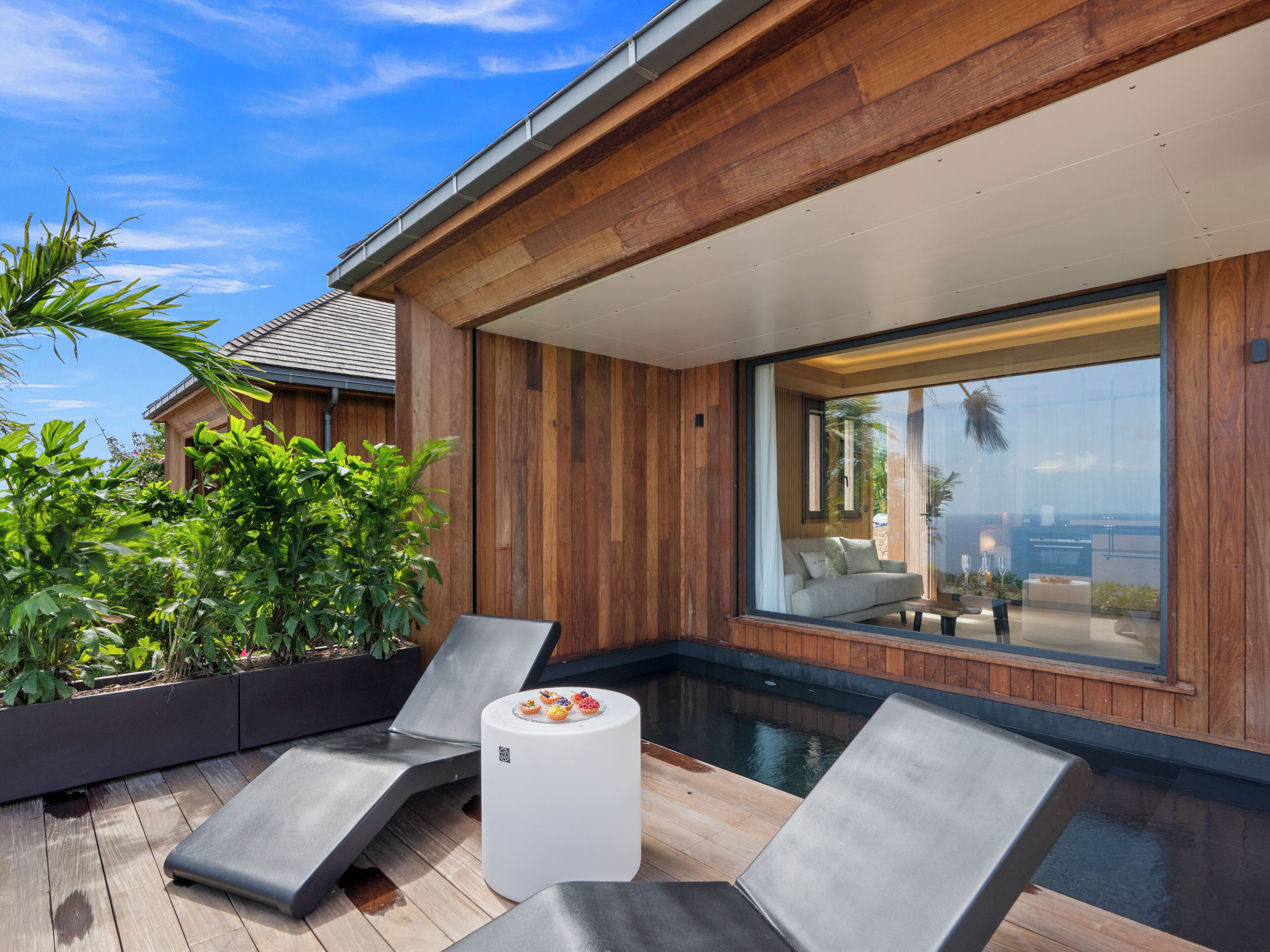 A modern outdoor pool area with lounge chairs and a small table, surrounded by wooden paneling and large windows offering views of the sky and a distant landscape.