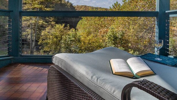 Une pièce avec un lit, un livre ouvert et une vue sur les arbres et le ciel, visible à travers une grande fenêtre.