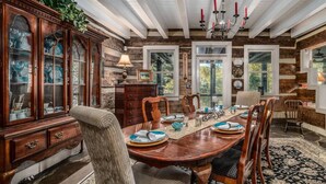 Une salle à manger avec une table en bois dressée pour six personnes, entourée de chaises. Un grand buffet en bois, doté de portes en verre, se trouve à gauche, et un lustre est suspendu au plafond.