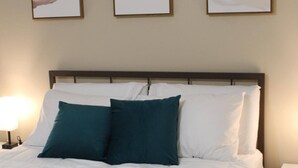 A neatly made bed with white bedding and teal pillows, flanked by two bedside lamps, in a room with three abstract paintings on the wall.