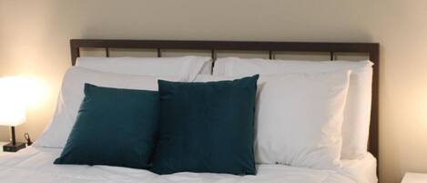 A neatly made bed with white bedding and teal pillows, flanked by two bedside lamps, in a room with three abstract paintings on the wall.