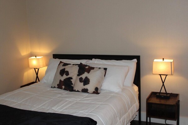 Un lit bien fait, avec une literie blanche et un oreiller à motifs de vaches, flanqué de deux tables de chevet sur lesquelles sont posées des lampes.