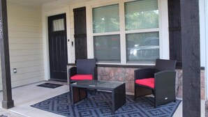 A porch with two wicker chairs with red cushions, a glass-top table, and a black door.