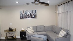 A living room with a grey sectional sofa, a small ottoman, a wall-mounted ceiling fan, and a cityscape painting.