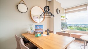 Ein moderner Essbereich mit Holztisch, Stühlen, einer hängenden Pendelleuchte, einem runden Spiegel und Blick ins Grüne durch ein Fenster.