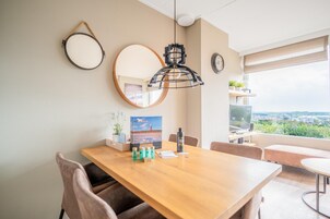 Ein moderner Essbereich mit Holztisch, Stühlen, einer hängenden Pendelleuchte, einem runden Spiegel und Blick ins Grüne durch ein Fenster.