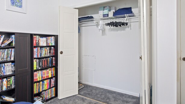 Um quarto com uma estante repleta de livros, um guarda-roupa com roupas penduradas e uma porta que dá para outro cômodo.