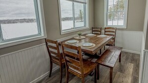 A dining room with a wooden table and chairs, large windows with blinds, and a view of a snowy landscape. - Lakeside Lodge | Waterfront | Private Dock (Hammond)