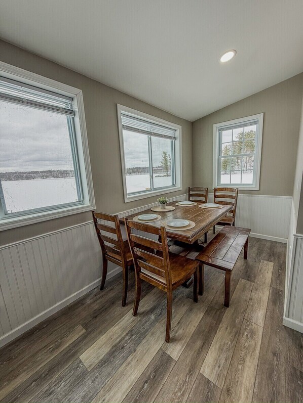 Une salle à manger avec une table et des chaises en bois, de grandes fenêtres équipées de stores et une vue sur un paysage enneigé.