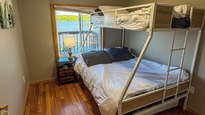 A bunk bed with a window view, a bedside table with a lamp, and a wooden floor.