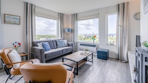 Un salon moderne avec un canapé gris, un fauteuil en cuir marron, une table basse en bois et de grandes fenêtres donnant sur le paysage.