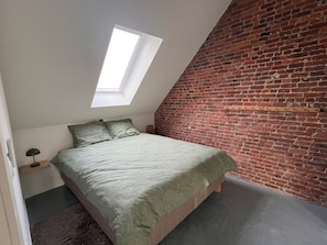 A bedroom with a sloped ceiling, a skylight, a brick wall, a bed with green bedding, and a small bedside table with a lamp. - Inviting Apartment in Bree With Terrace (Bree)