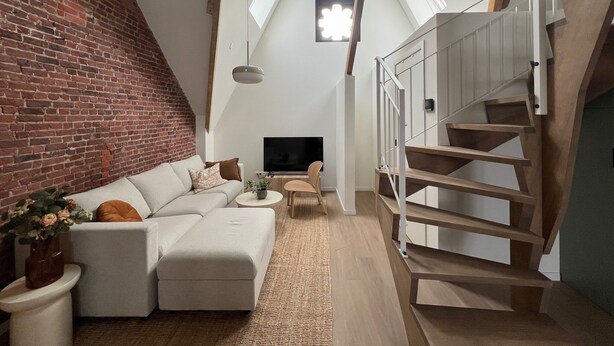 Un ambiente living in stile loft con parete in mattoni, un divano bianco, una televisione, una scala e lucernari.