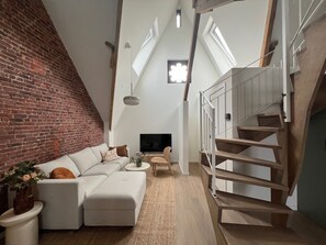 A loft-style living space with a brick wall, a white sofa, a TV, a staircase, and skylights. - Inviting Apartment in Bree With Terrace (Bree)