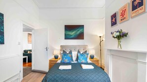 Una habitación de hotel con una cama, dos mesitas de noche, una obra de arte en la pared, un jarrón con flores y un pasillo que conduce a otra habitación.