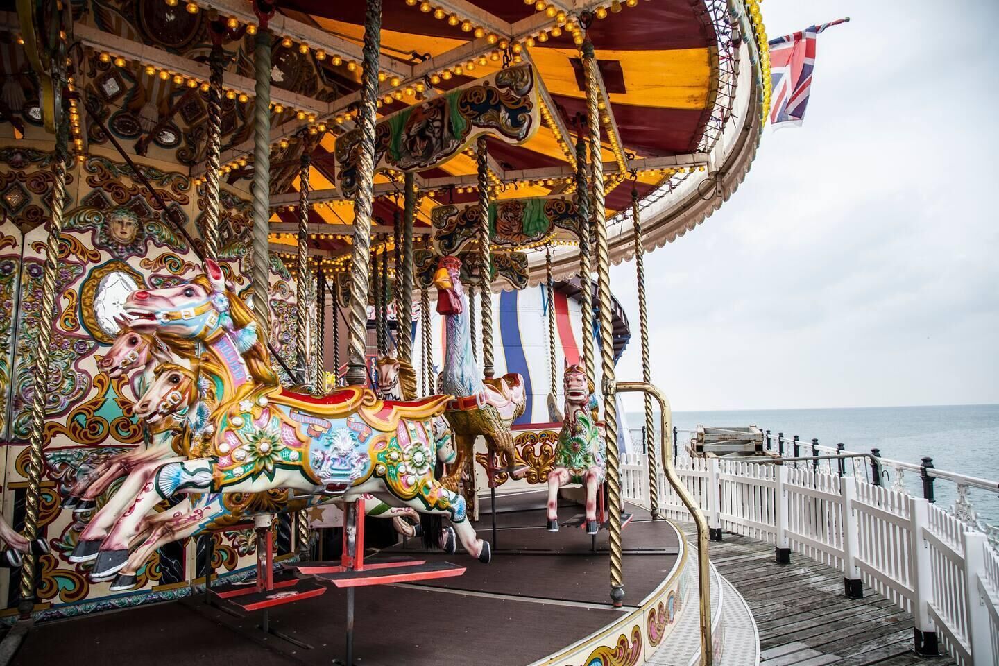 A carousel with ornate horses and a flag, located on a pier with a view of the sea.